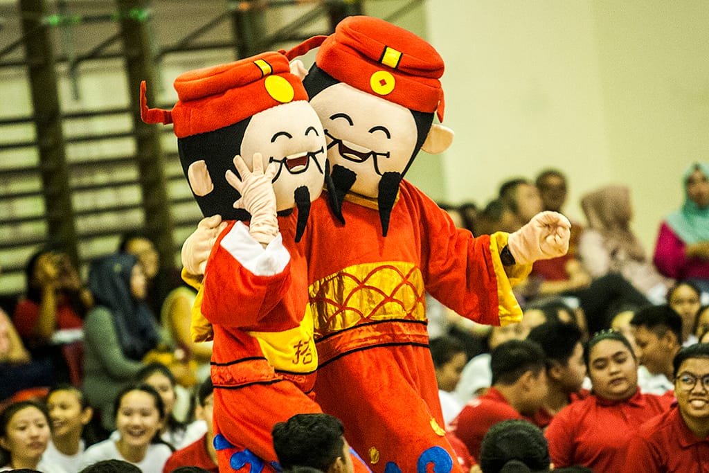 Two smiling Chinese deity mascots in red and gold costumes perform at a crowded indoor event.